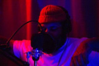 An independent rapper standing in a dimly lit studio booth performing into a condenser microphone.