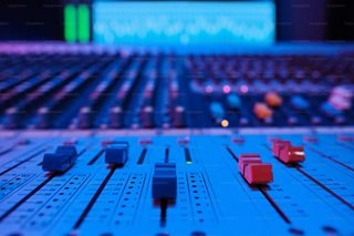 A close-up horizontal perspective of a professional audio mixing console and studio speakers glowing inside a modern music studio.