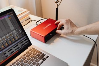 Hand adjusting the volume knob on a red Focusrite audio interface while auditioning beat waveforms on a laptop.